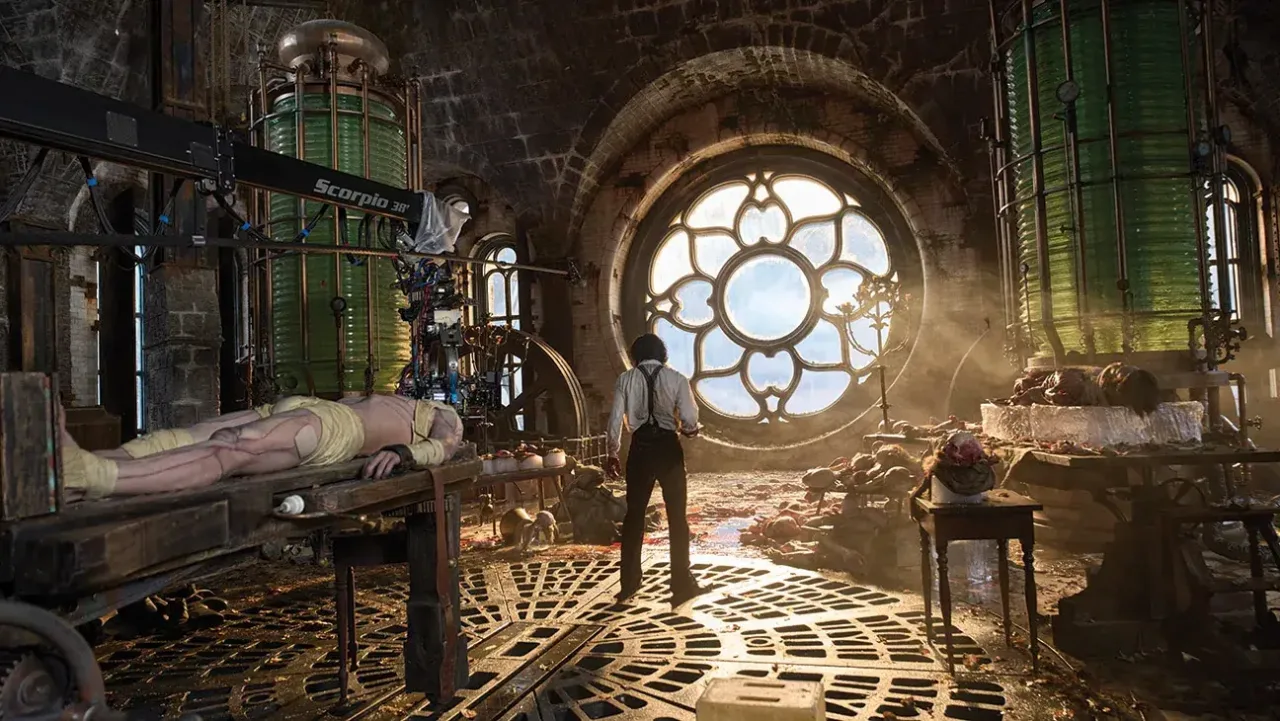 A behind-the-scenes production photograph capturing a sprawling, detailed laboratory set with a Gothic architectural aesthetic. At the center, a man stands facing away from the camera, wearing a light-colored button-up shirt with dark suspenders and dark trousers. He stands on a floor made of heavy, concentric circular iron grates, looking toward a massive, ornate circular rose window that dominates the back wall and looks out into a bright blue sky. To his left, a large, draped biological specimen or figure lies horizontally across a sturdy wooden operating table, with a prominent black mechanical camera crane labeled "Scorpio 38" extended directly above it. On the right side of the room, massive cylindrical glass fermentation tanks filled with translucent green fluid rise toward the arched brick ceiling, surrounded by metal framework, piping, smaller tables covered with anatomical specimens, and jars. Soft, atmospheric golden sunlight pours through the central window, casting a warm haze over the entire industrial space.